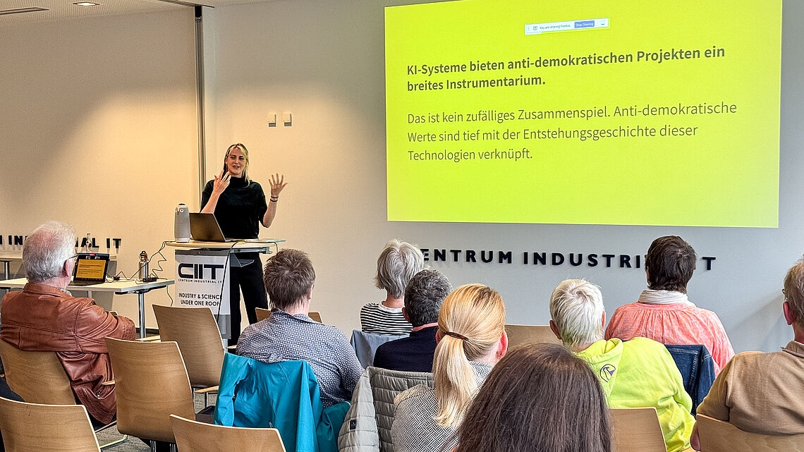 Annemarie Witschas steht vor dem Publikum und spricht neben einer Präsentationsfolie über anti-demokratische Projekte und KI-Systeme.